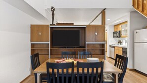 A compact kitchen with wooden cabinets, a built-in TV, and a dining area with a table and chairs. - The Traverse by Avantstay Ski-in/ski-out Condo Pool, Hot Tub Access Steps to Kt-22 (Olympic Valley)