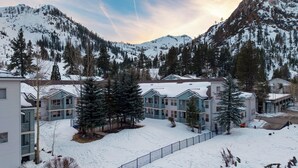 A snow-covered mountain resort with multiple-story buildings, a fenced pathway, and a clear view of the surrounding mountains. - The Traverse by Avantstay Ski-in/ski-out Condo Pool, Hot Tub Access Steps to Kt-22 (Olympic Valley)