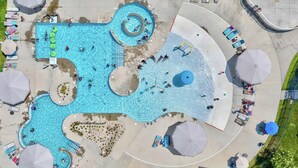 En stor, uregelmæssigt formet swimmingpool med flere afdelinger og en ø i midten, omgivet af liggestole og parasoller.