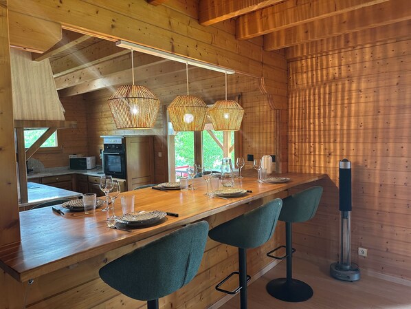 L’intérieur d’un chalet en bois comprend une longue table à manger, des tabourets de bar, des lampes suspendues en osier et un coin cuisine.