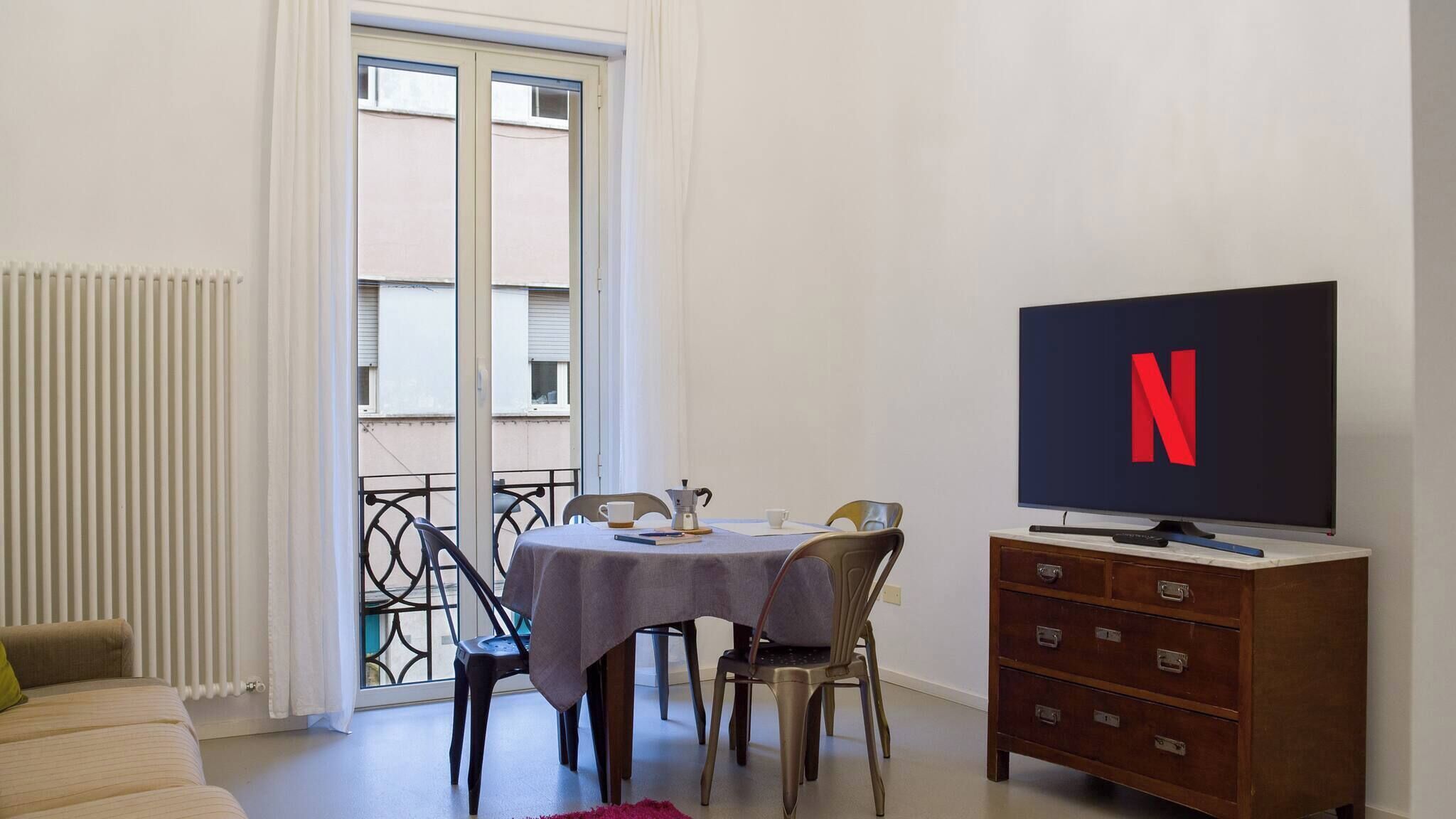 Una stanza con un tavolo, sedie, una TV con il logo Netflix, un termosifone e una finestra con vista.