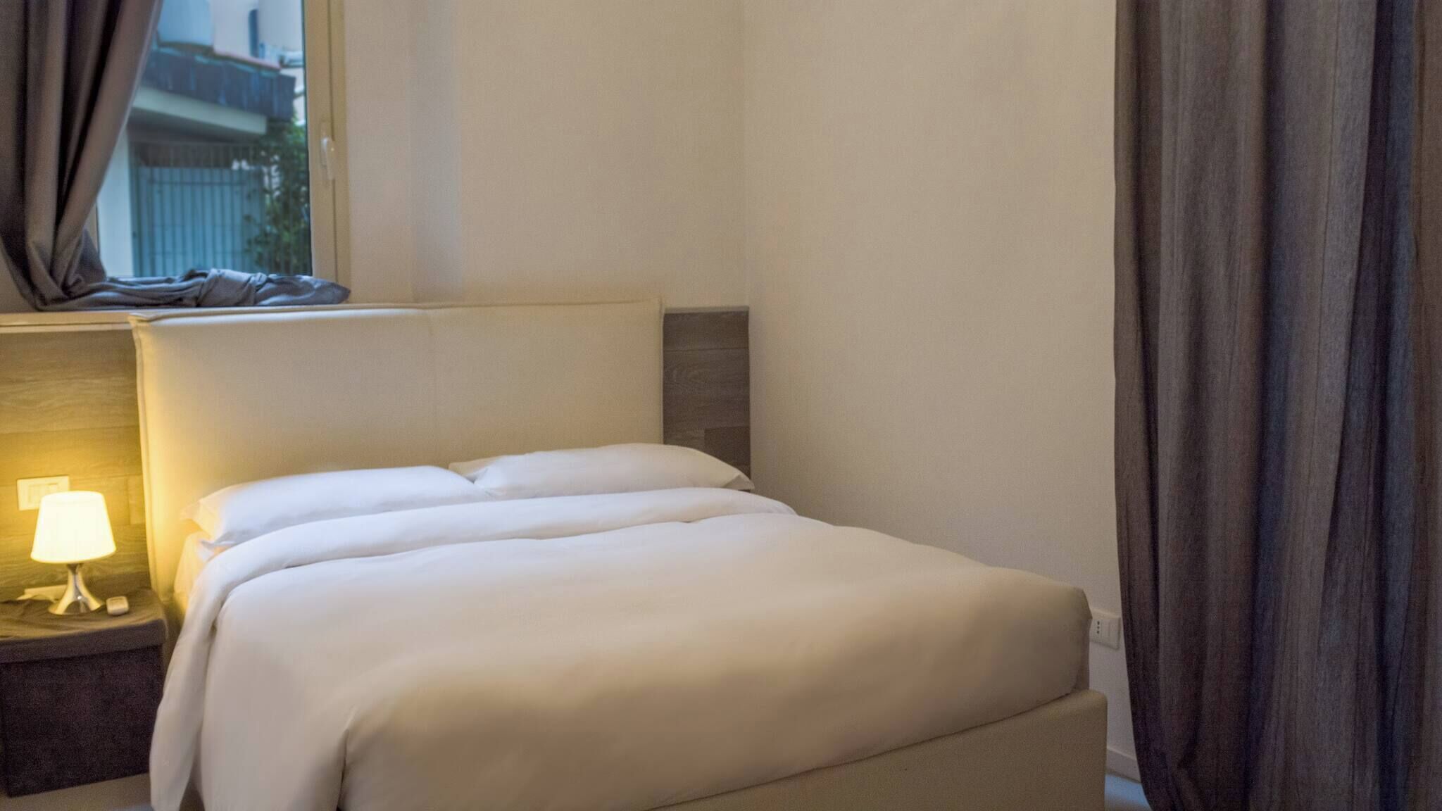 Siististi pedattu sänky hotellihuoneessa, jossa on yöpöytälamppu, sängynpääty ja ikkuna, jossa on verhot.