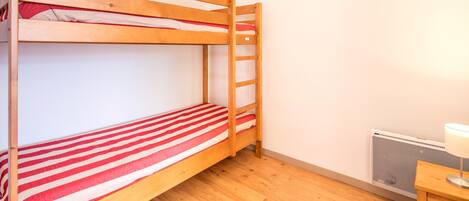 Un lit superposé avec une literie rouge et blanche, une échelle, une lampe fixée au mur et un radiateur.