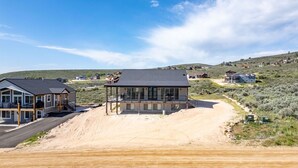 A modern house with a covered porch, a driveway, and a hilly landscape in the background. - The Lazy Days Getaway at Bear Lake with Ideal Beach Access (Garden City)
