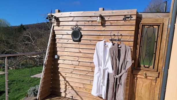 A wooden cabin with a hot tub, wooden walls, a door with a glass panel, and a coat rack with robes. - Gîte des Petites Pyrénées (AUSSEING)