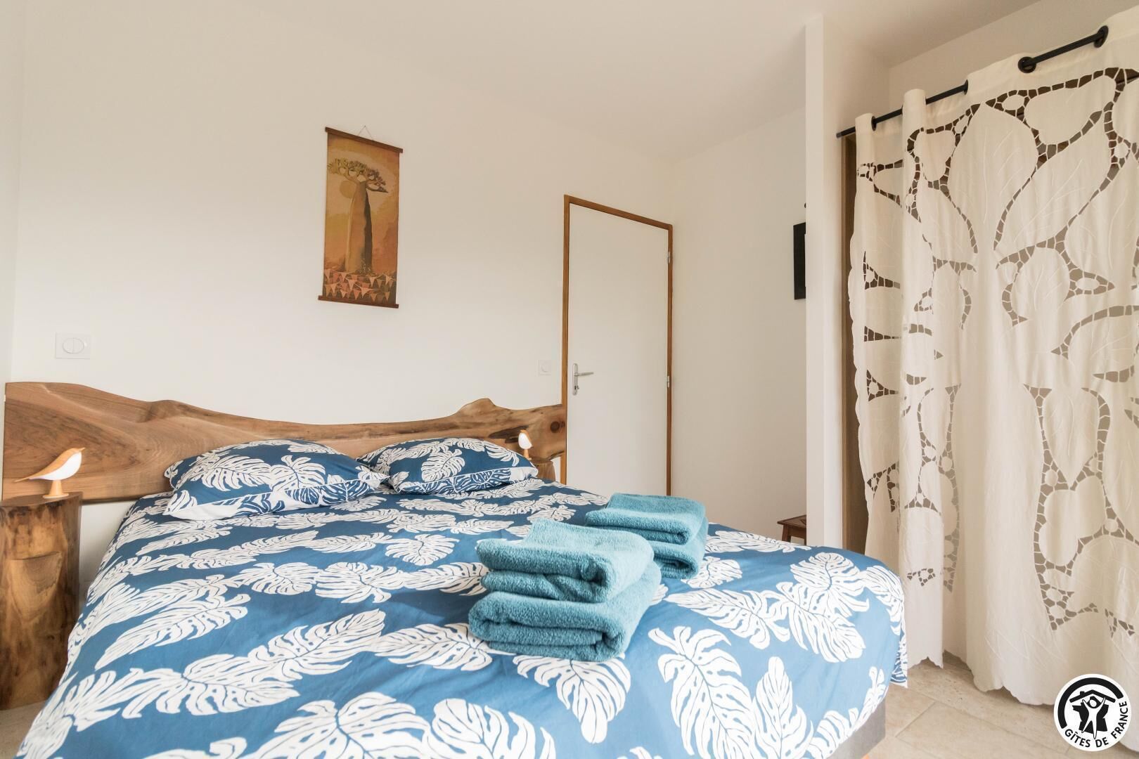 Ein Schlafzimmer mit Holz-Kopfteil, einem Bett mit blau-blumiger Bettdecke, zwei blauen Handtüchern, einem Nachttisch mit Lampe, einem gerahmten Bild an der Wand und einer Tür mit Spiegel.