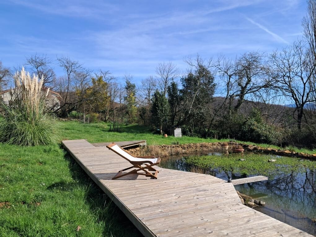 Une terrasse en bois avec un fauteuil de relaxation qui s’étend au-dessus d’un étang, le tout entouré de verdure et d’arbres.