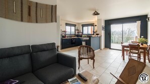 A modern living room with a sofa, a dining table with chairs, a kitchen area, and a balcony with a view. - Gîte des Petites Pyrénées (AUSSEING)