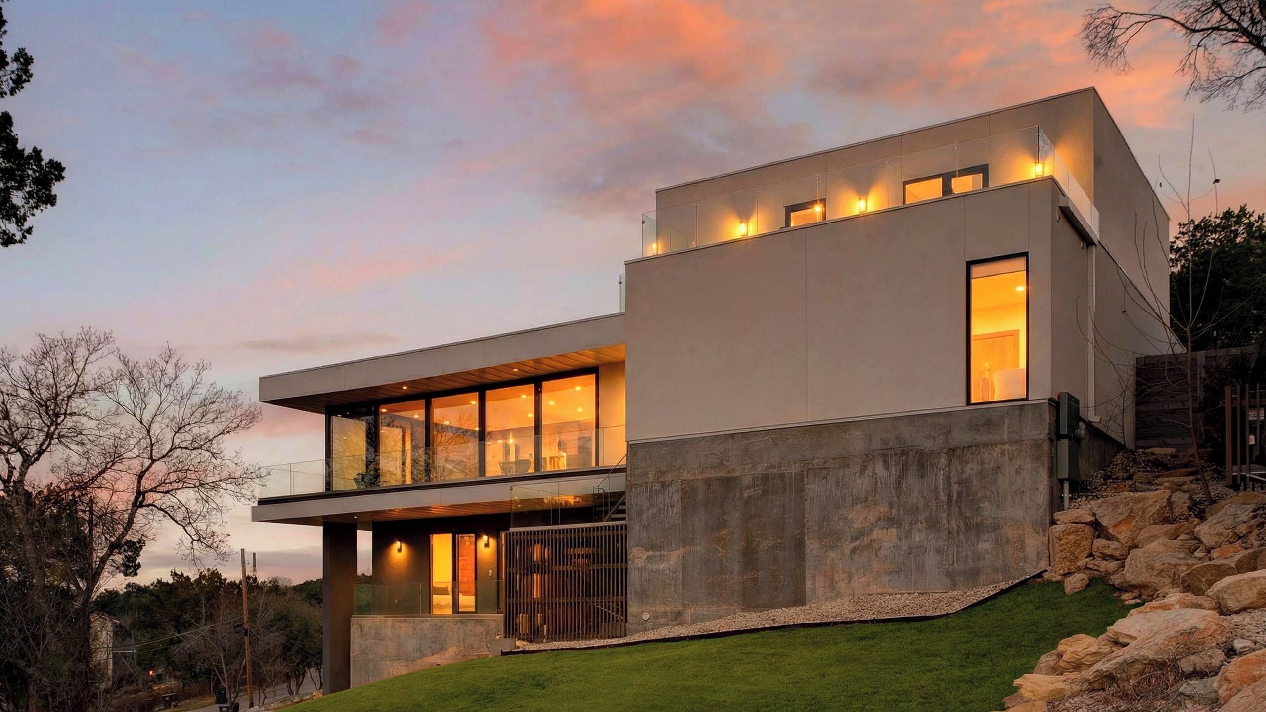 A modern two-story house with large windows, a flat roof, and a stone retaining wall.