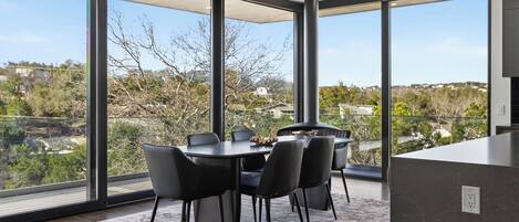 A modern dining area with a round table and chairs, large windows with a view of a residential area, and a kitchen counter.