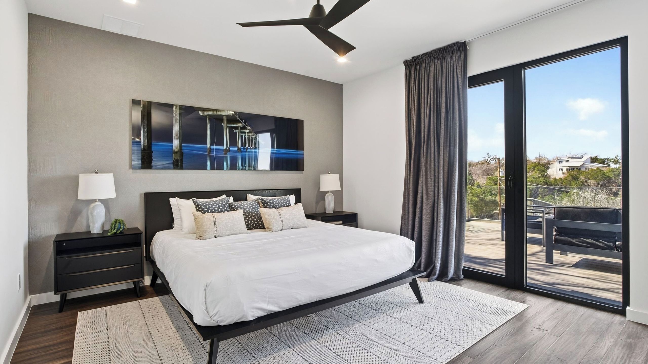 Ein modernes Schlafzimmer mit einem großen Bett, einem Deckenventilator, einem Gemälde und Blick durch ein großes Fenster.