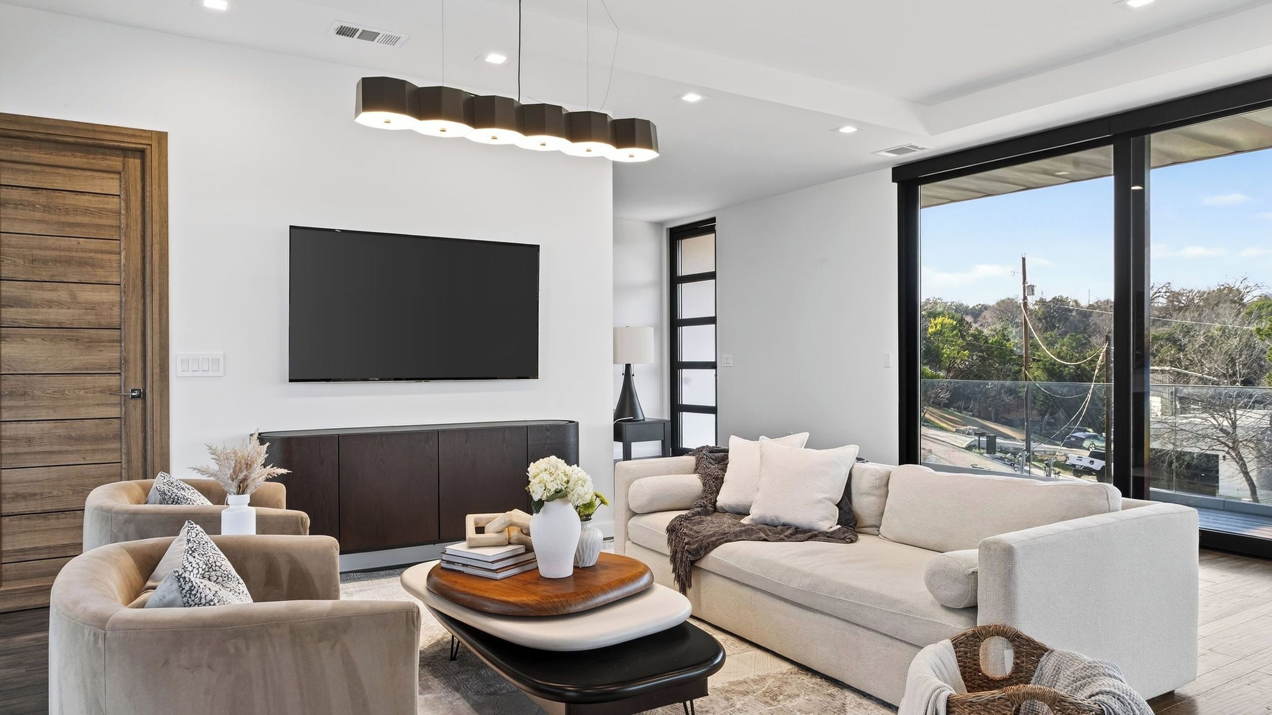 A modern living room with a flat-screen TV, comfortable seating, and a large window offering a city view.