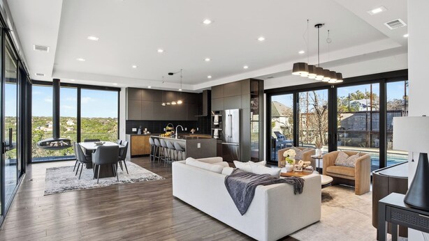 A modern living room with a sofa, a dining area with chairs, a kitchen with dark cabinets, and large windows offering a view of the outdoors. - Hunters Estate by Avantstay Design-forward Hillside Home Private Pool + Firepit Close to Lake Travis + Downtown Austin (Austin)