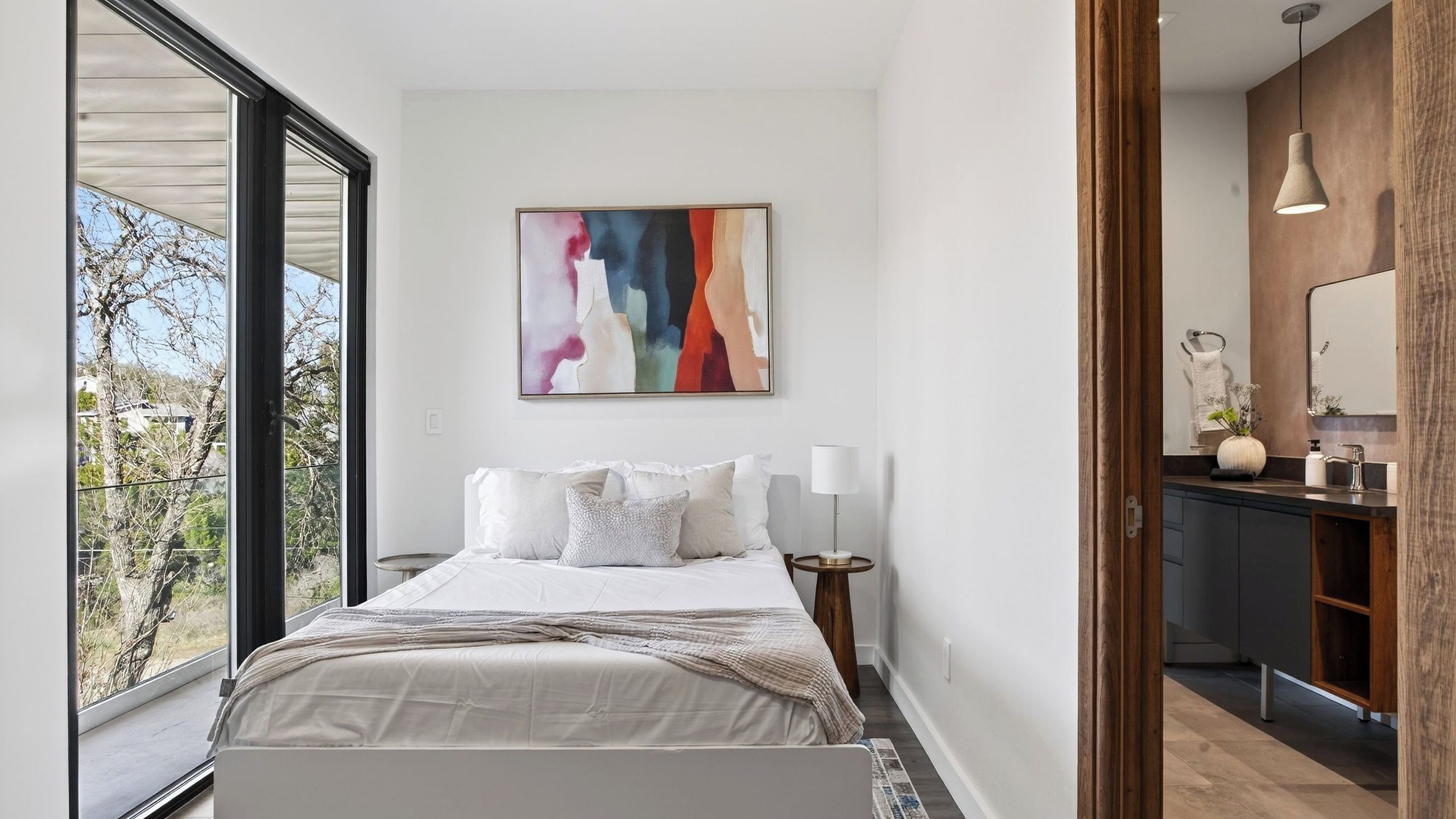 Une chambre moderne dotée d’une grande fenêtre, d’un lit avec du linge de lit blanc, d’un tableau abstrait aux couleurs vives et d’une salle de bain avec un meuble-lavabo en bois.