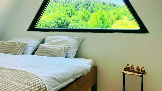 Quarto com cama de madeira, roupa de cama branca, uma pequena mesa de cabeceira com potes e vista para uma floresta por uma janela triangular.