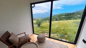 Quarto com grande janela que oferece vista para uma paisagem verde e montanhas. Há uma cadeira com almofadas e uma pequena mesa redonda.