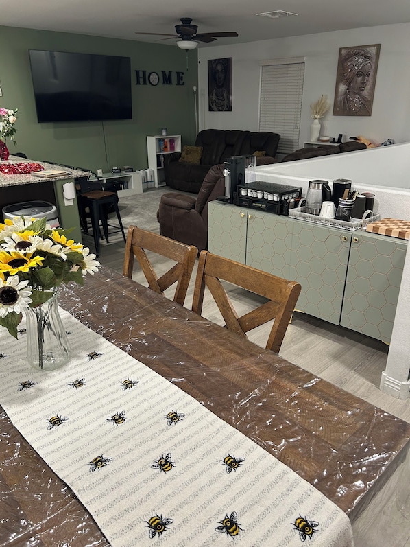 Une cuisine moderne avec un coin repas, comprenant un îlot de cuisine, une table à manger recouverte d’une housse de protection et un vase rempli de tournesols.