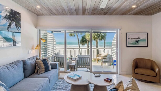 A modern living room with a large window overlooking a beach, a ceiling fan, and a sofa with cushions. - Beachfront Bliss - Outdoor Pool - Sleeps 6 (Belleair Beach)