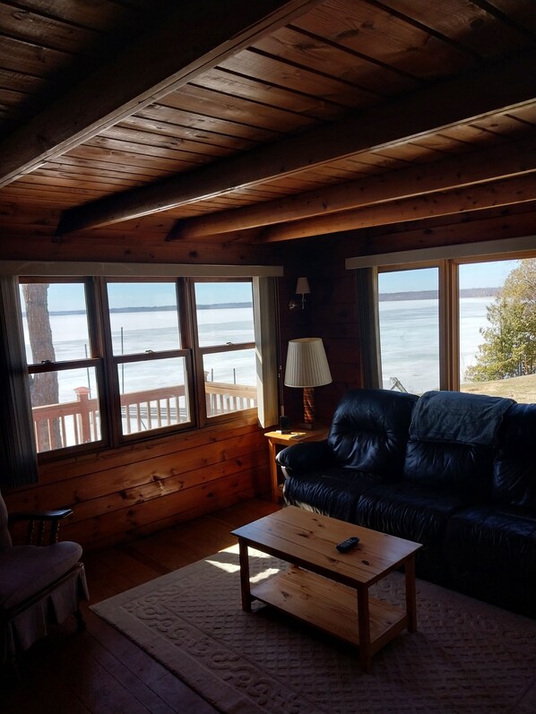 Eine Holzhütte mit einem großen Fenster mit Blick auf ein Gewässer, einem Ledersofa, einem Holztisch und einer Lampe.