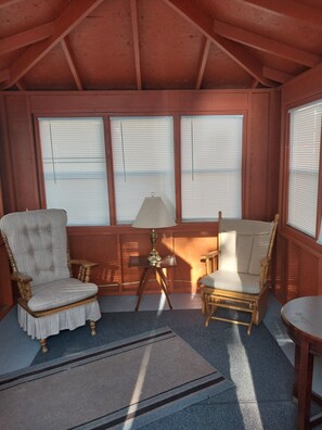 A cozy room with wooden beams, a large window with blinds, a lamp on a small table, and two upholstered chairs.