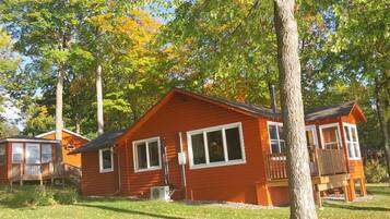 Une cabane avec une terrasse, un petit porche et un grand arbre à l’avant.