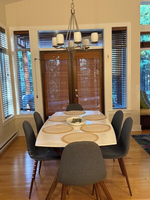 A dining room with a large table, six chairs, a chandelier, and large windows with blinds.