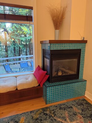 A cozy living room with a fireplace, a bench with cushions, and a view of a forest through a large window.