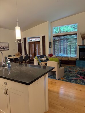 A modern kitchen with a black countertop, a dining area with chairs and a table, a sofa with colorful cushions, and a large window with blinds.