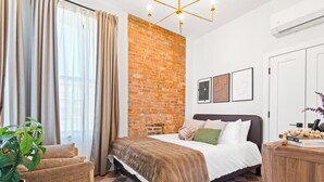 A bedroom with a brick wall, a bed with a brown blanket, a wooden nightstand, a potted plant, and a checkered floor. - The Bergen Standard  3BD 2BA Brownstone + Parking (Jersey City)
