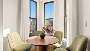 A round wooden table with a vase and plant, surrounded by four green upholstered chairs in a room with large windows and white curtains. - The Bergen Standard  3BD 2BA Brownstone + Parking (Jersey City)