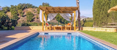 A swimming pool with a pergola and seating area, surrounded by greenery and hills.