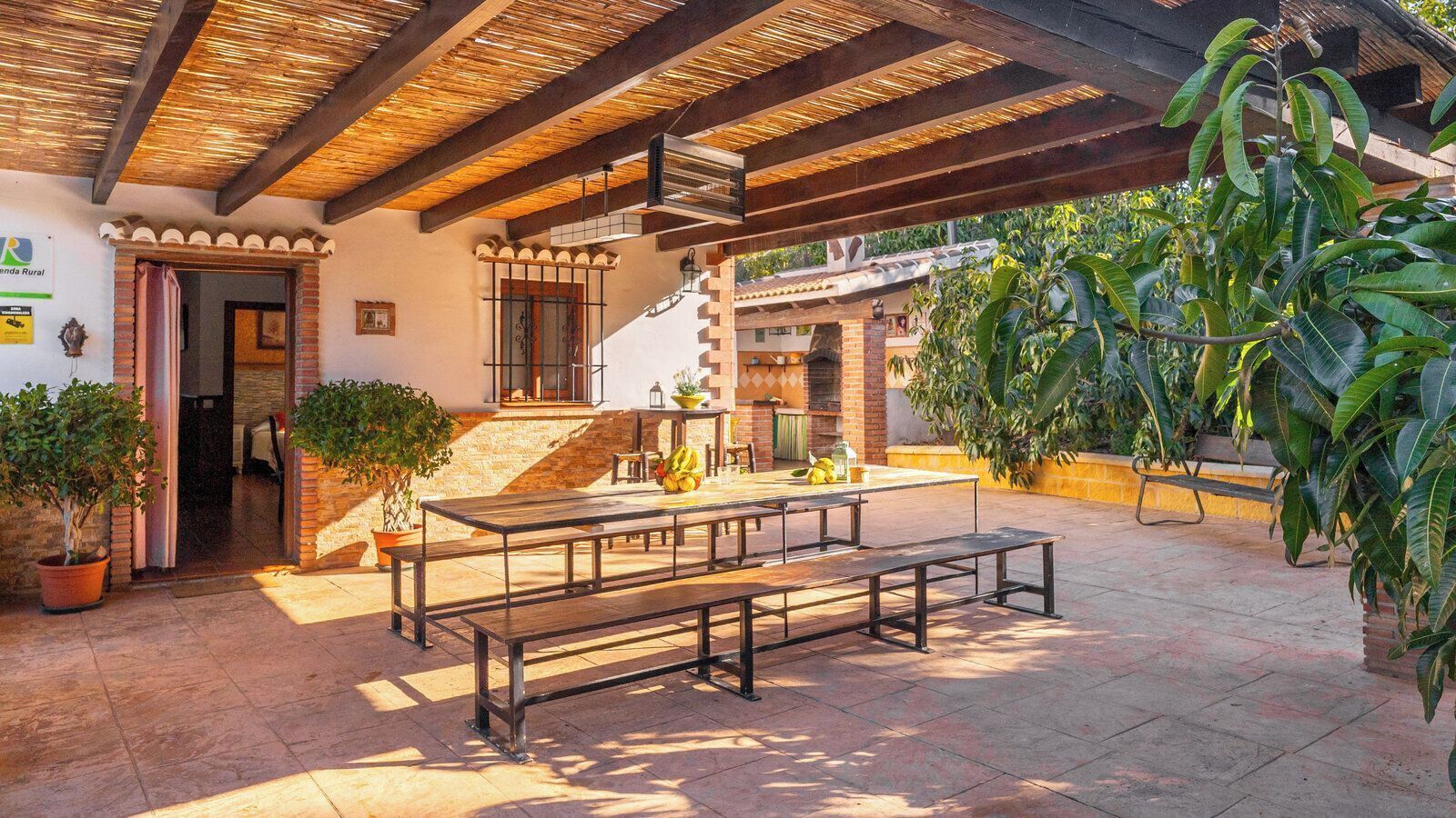 An outdoor patio with a wooden table and benches, surrounded by potted plants and a brick wall.
