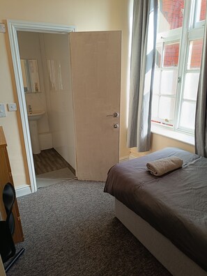 Quarto com cama, porta para um banheiro, janela com cortinas e piso coberto por carpete.