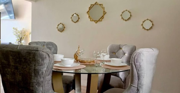 Uma área de refeições com uma mesa redonda de vidro, cadeiras estofadas cinzas, pratos brancos e espelhos decorativos dourados na parede.