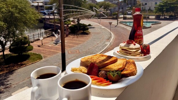 A breakfast setting with toast, pancakes, and fruit on a balcony overlooking a street with buildings and a pool. - ARENA DORADA (Puerto Baquerizo Moreno)