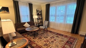 A living room with a wooden floor, a round coffee table, a sofa, a chair, a lamp, a clock, and a window with curtains. - Peaceful 1-bedroom apartment with AC in vibrant Minneapolis (Minneapolis)
