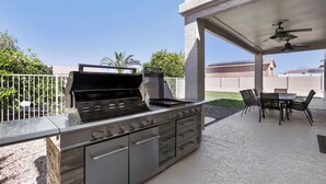 Een terras buiten met een ingebouwde barbecue, een tafel met stoelen en een plafondventilator.