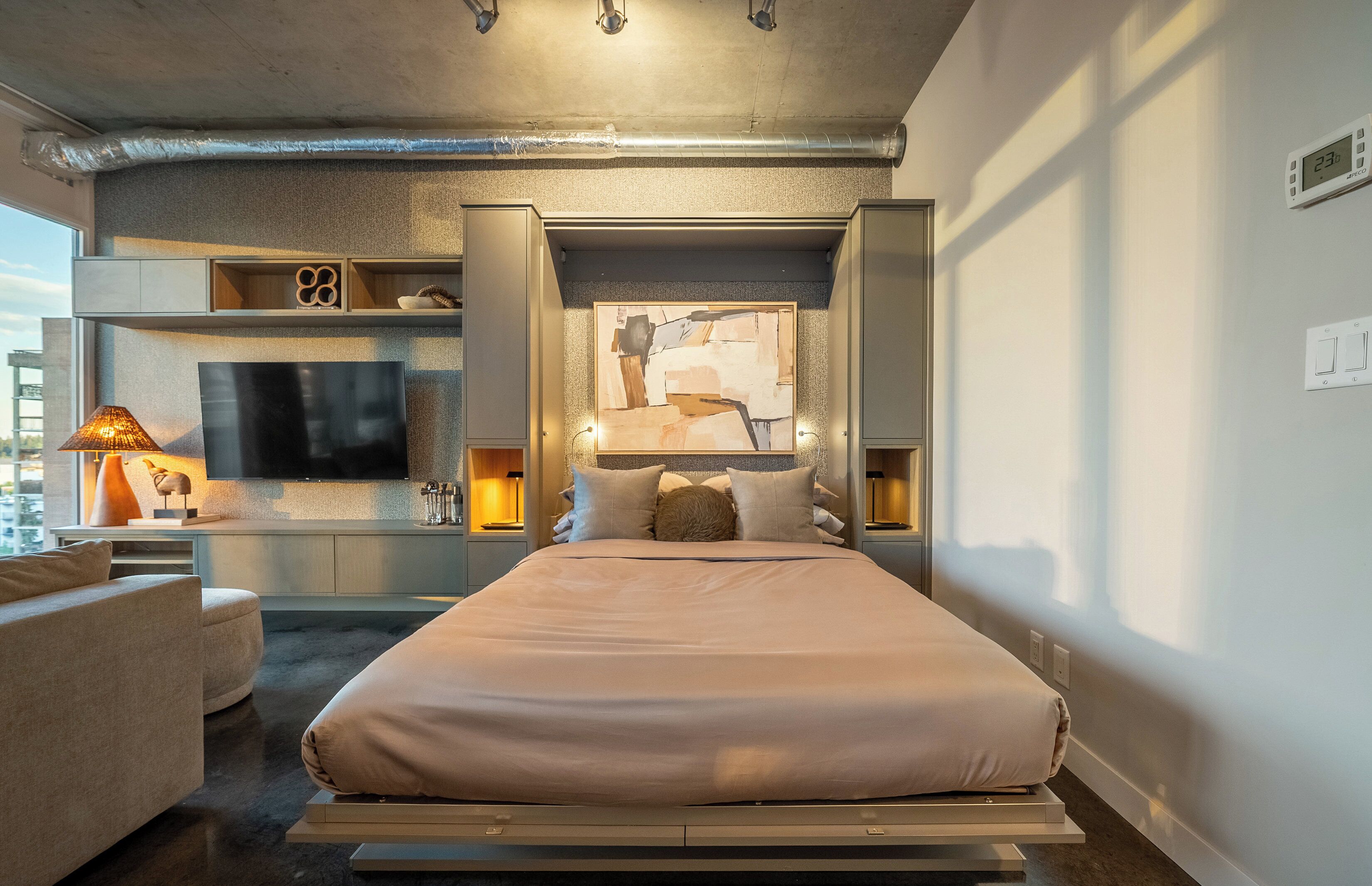 A modern bedroom with a large bed, a flat-screen TV, built-in shelving, and a city view.