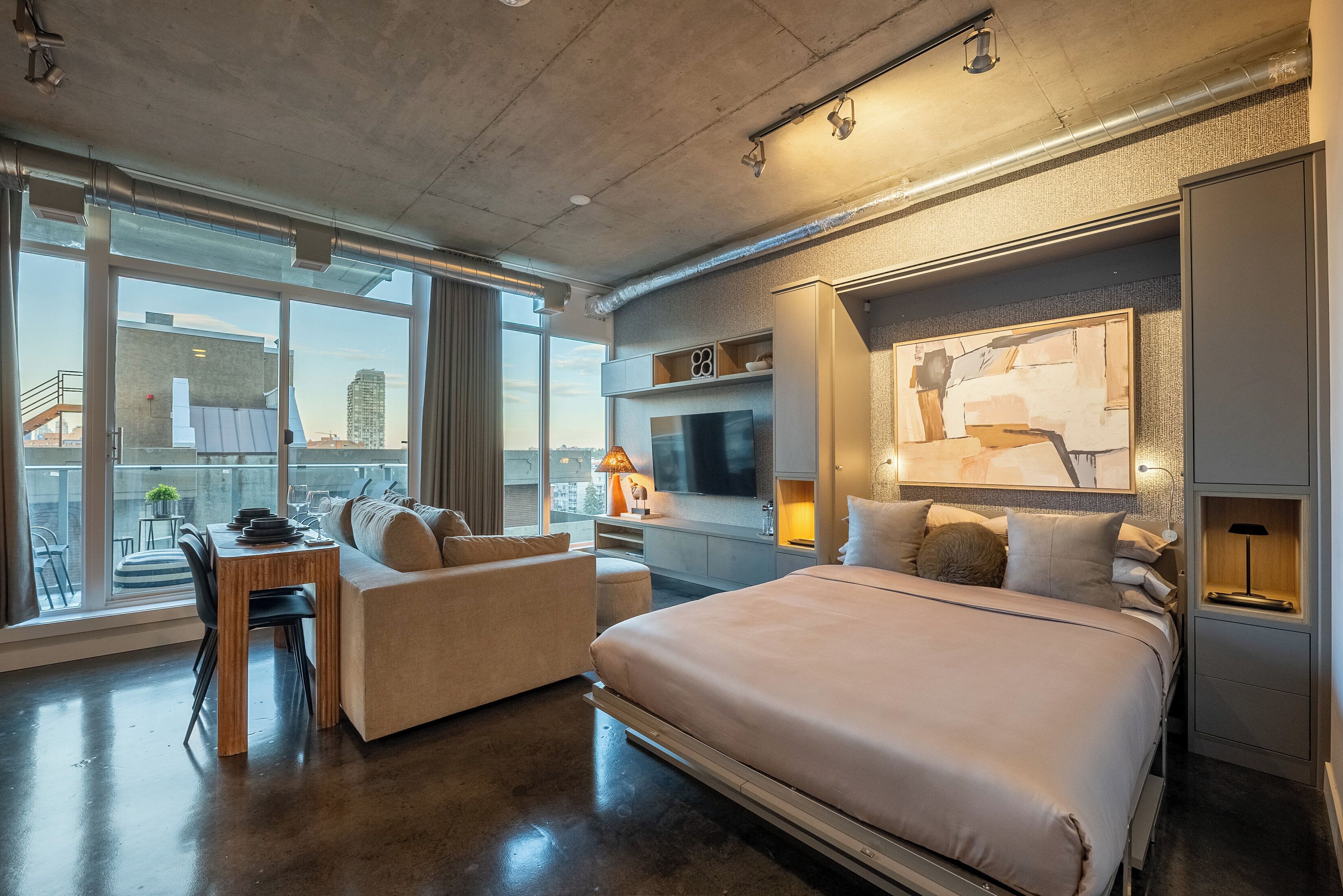 A modern hotel room with a large bed, a sofa, a dining table, and a TV.