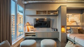 Ein modernes Hotelzimmer mit großem Fenster, einem an der Wand befestigten Flachbildfernseher, einem Bett mit Kopfteil und einer Sitzecke mit Hocker.