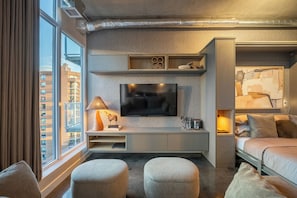 A modern hotel room with a large window, a flat-screen TV mounted on a wall, a bed with a headboard, and a seating area with ottomans.