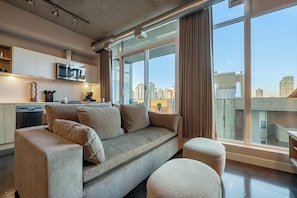 A modern living room with a sofa, ottoman, and a large window offering a city view.