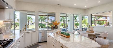 A modern kitchen with a large island, white cabinetry, and a view of a landscaped outdoor area.