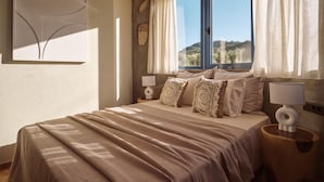 Une chambre à coucher avec un grand lit, une fenêtre avec des rideaux, une lampe fixée au mur et une table de chevet en bois.