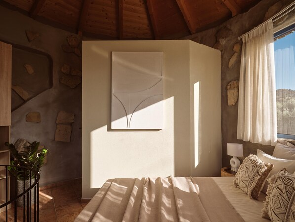 Un dormitorio con cama, una obra de arte abstracta en la pared, planta en maceta y vistas al exterior a través de una ventana.