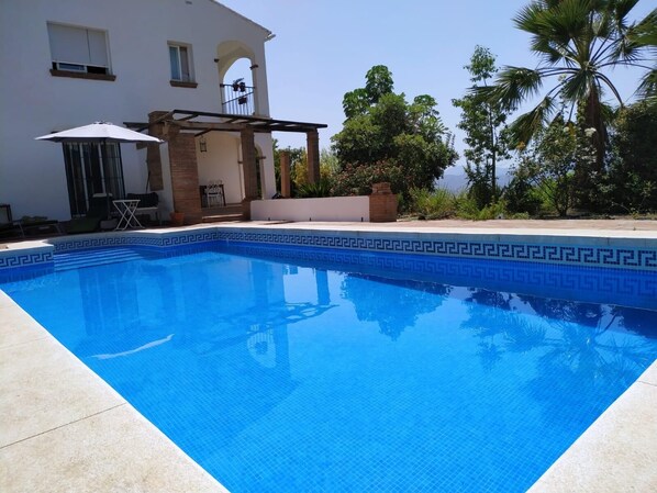 A swimming pool with a blue mosaic border, a patio area with a table and chairs, and a white building with arched windows.