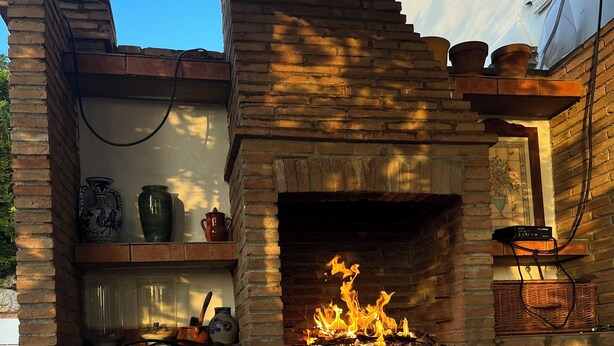 A brick-built outdoor fireplace with a fire burning inside, surrounded by shelves with decorative items and a bench. - Villa La Malcontenta, large private house with green spaces and swimming pool. (Alcaucín)