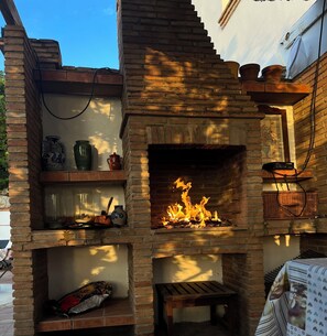 A brick-built outdoor fireplace with a fire burning inside, surrounded by shelves with decorative items and a bench.