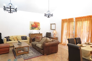A living room with a sofa, coffee table, and dining table set for two. There are two chandeliers, a painting, and a lamp on a side table.