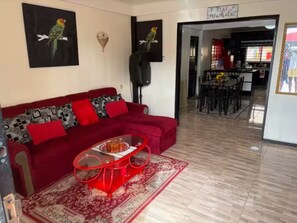 A living room with a red sofa, a glass coffee table, and a red rug. There are two wall-mounted speakers and a dining area in the background. - 3-bedroom house in quaint Paramaribo with cool AC breeze. (Paramaribo)